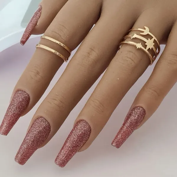 A hand with long, glittery pink coffin-shaped made-to-order press-on nails wears two gold rings—one with parallel lines, the other featuring star and moon cutouts.