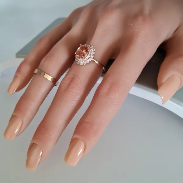 A hand with manicured nude, made to order press-on nails wears two rings—one plain gold band and one ornate ring with a large, oval gemstone surrounded by small diamonds. The hand rests on a light surface.