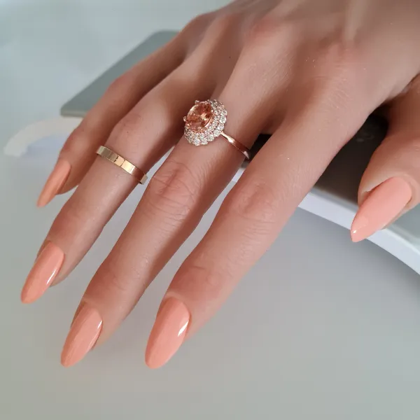 A hand with long, peach-colored made to order press-on nails wears two rings: a plain gold band on the middle finger and a diamond-encrusted ring with a large central stone on the ring finger.