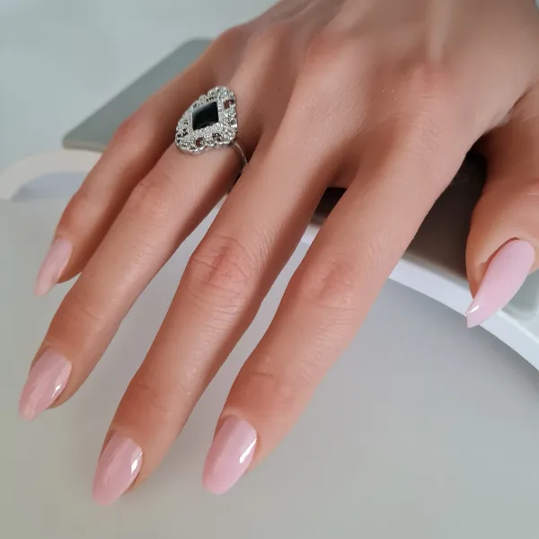 A hand with long, pale pink made to order press-on nails wears a large, ornate ring featuring a black center stone surrounded by clear stones. The hand rests on a white and gray surface.