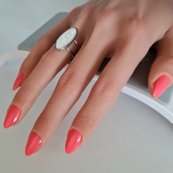 A hand with bright coral pink, almond-shaped made to order press-on nails and a large oval ring with a white stone rests on a flat surface.
