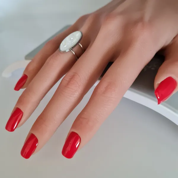 A hand with neatly manicured, bright red made to order press-on nails rests on a white surface. The person is wearing a silver ring with a large, oval white stone on their middle finger.