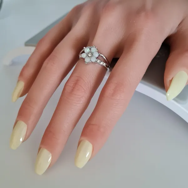 A hand with long, pale yellow made to order press-on nails wears a silver floral ring with light blue stones and small diamonds on the ring finger.