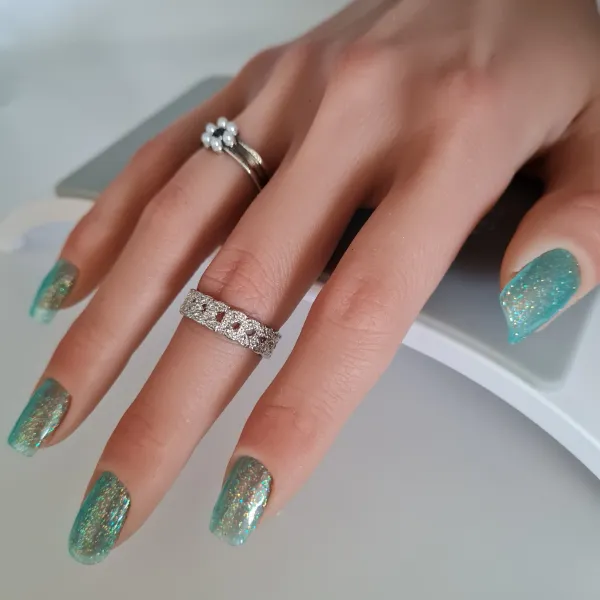 A hand with long, sparkly turquoise made to order press-on nails wears two silver rings: one with a flower design and one with embedded gems. The hand rests on a white and gray surface.