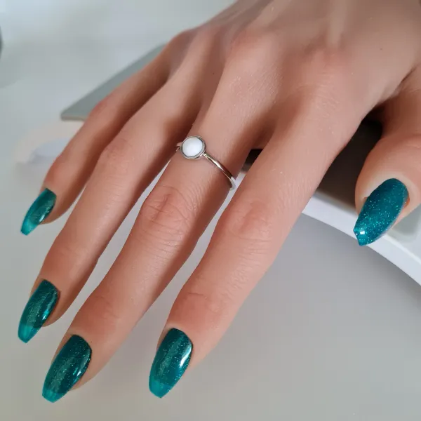 A hand with teal, glittery made to order press-on nails and an oval white stone ring rests on a white surface.