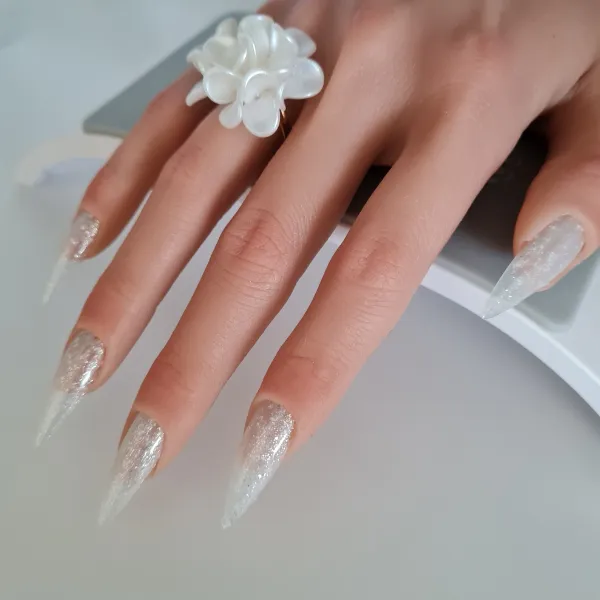A hand with long, pointed, glittery silver made to order press-on nails rests on a white surface. The person is wearing a large, white, flower-shaped ring on one finger.