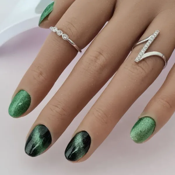 Hand with almond-shaped, made-to-order press-on nails, featuring a metallic green and black gradient polish, adorned with two silver rings: one set with small gemstones, the other in a distinctive V-shaped style.