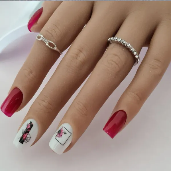 A hand with long, square, made-to-order press-on nails; some are red, others white with floral and geometric art. The hand wears two silver rings.