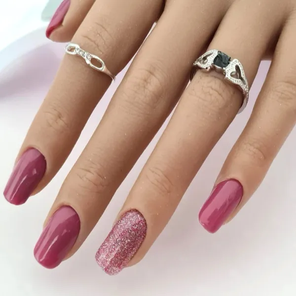 A hand with long, pink made-to-order press-on nails; the ring finger has a sparkly glitter polish. Two silver rings with different designs adorn the hand.