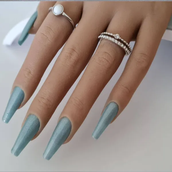 A hand with long, square-shaped, light blue press-on nails wears a pearl ring on the index finger and a stacked diamond ring on the ring finger against a plain background.