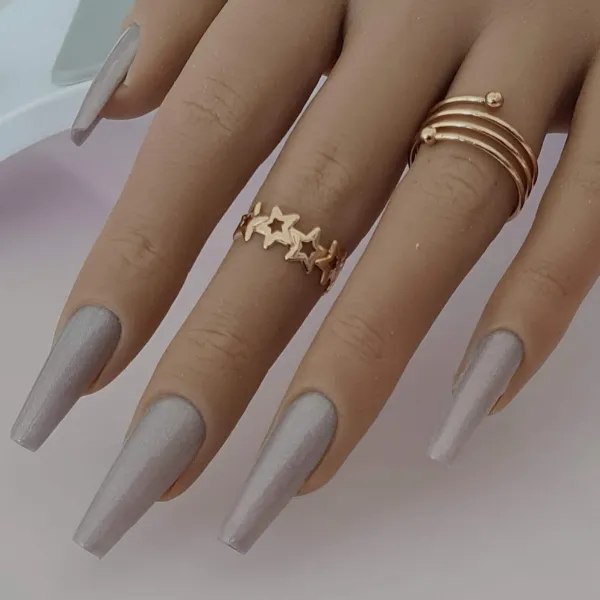 A hand with long, silver-gray coffin-shaped press-on nails wears two gold rings—one star-detailed and one with three wrapped bands and a bead—set against a soft, blurred background.