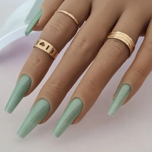 A hand shows off long coffin-shaped, light green made-to-order press-on nails, complemented by three gold rings of assorted designs.