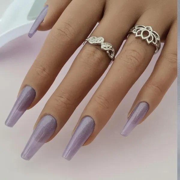 A hand wears long, coffin-shaped, shimmery lavender made-to-order press-on nails and two silver rings with decorative designs on the middle and ring fingers.