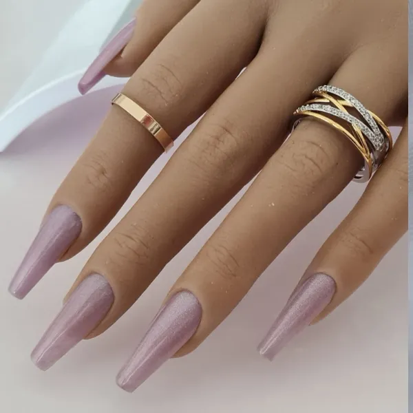 A hand with long, coffin-shaped, shimmery light purple made-to-order press-on nails. The fingers wear three gold and silver rings, set against a soft, light background.
