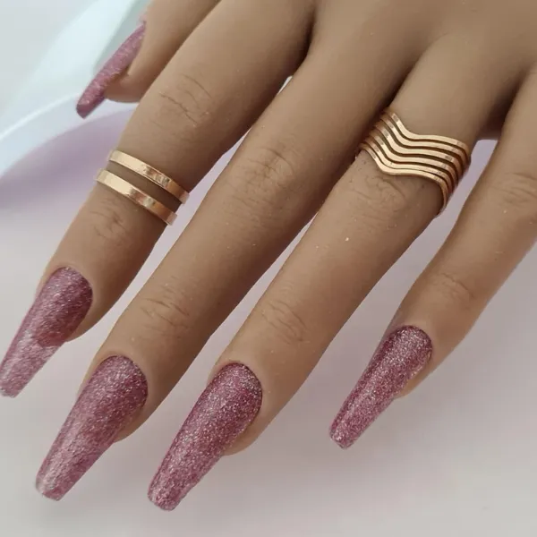 A hand with long, sparkly pink made-to-order press-on nails wears two gold rings—one with parallel bands on the middle finger and a wavy ring on the ring finger—against a light background.