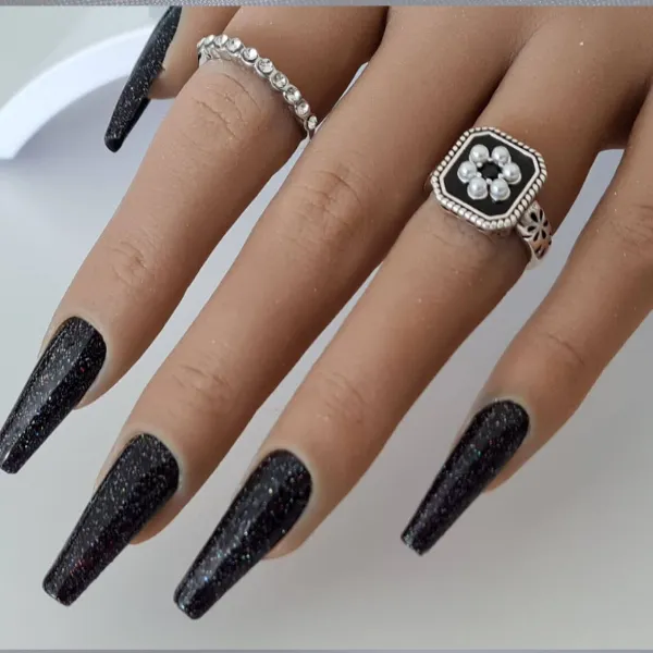 A hand with long, black glittery made-to-order press-on nails wears two silver rings—one floral, one with round beads. The fingers are gracefully spread on a white surface.
