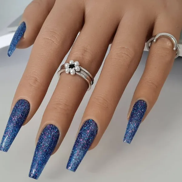 A hand displays long, blue glittery coffin-shaped made-to-order press-on nails and three silver rings—one featuring pearl accents—against a light background.