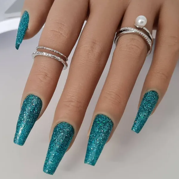 A hand with long, square-shaped nails in sparkly teal polish wearing silver rings, including a pearl ring. 23 press on nails are made to order for a stunning custom look.